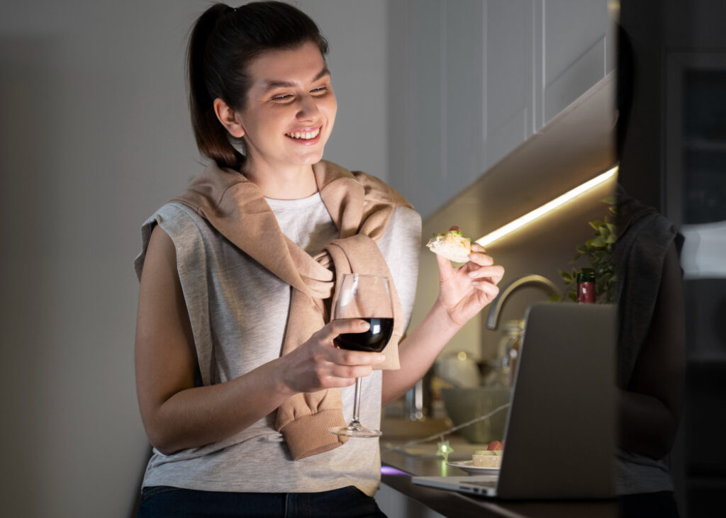 une femme mange et boit un verre de vin en souriant devant son ordinateur